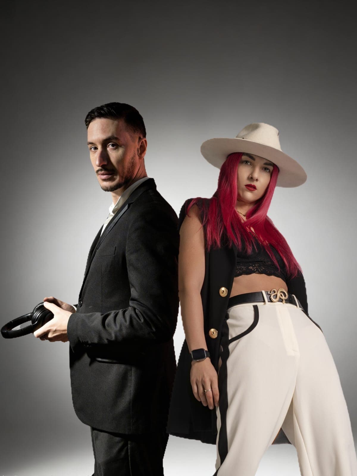 Uomo in abito scuro e donna con capelli rosa e cappello bianco schiena contro schiena.