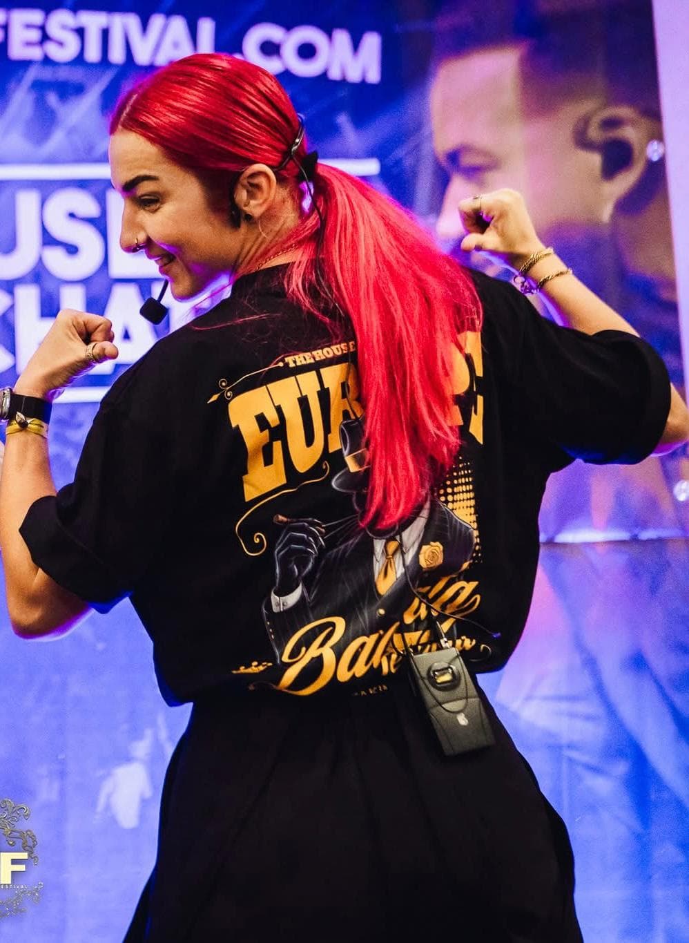 Donna con capelli rosso acceso flette i bicipiti indossando una maglietta nera con stampe.