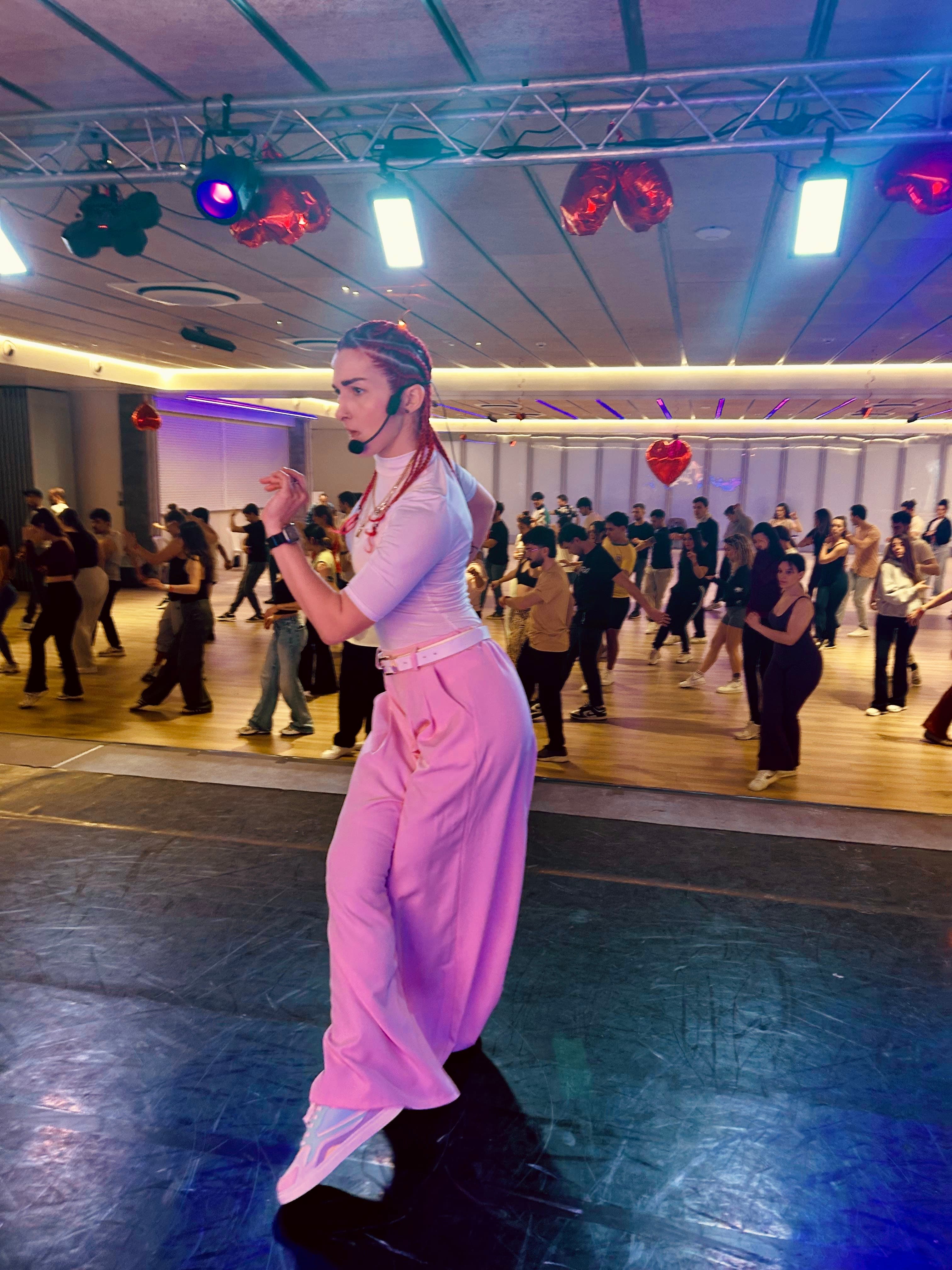 Donna con treccine rosse e pantaloni rosa guida una lezione di danza in uno studio.