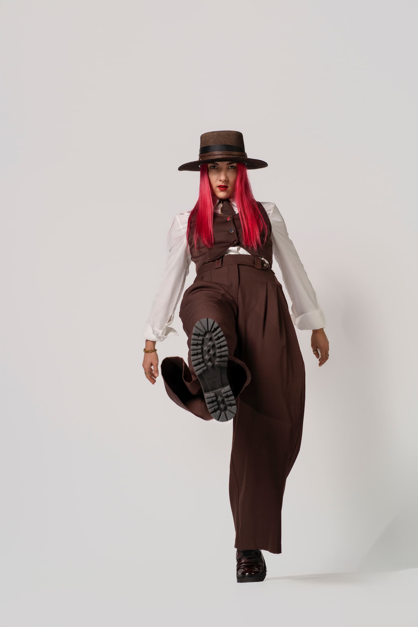 Donna con capelli rosa in abito marrone calcia verso l'obiettivo mostrando la suola dello stivale.