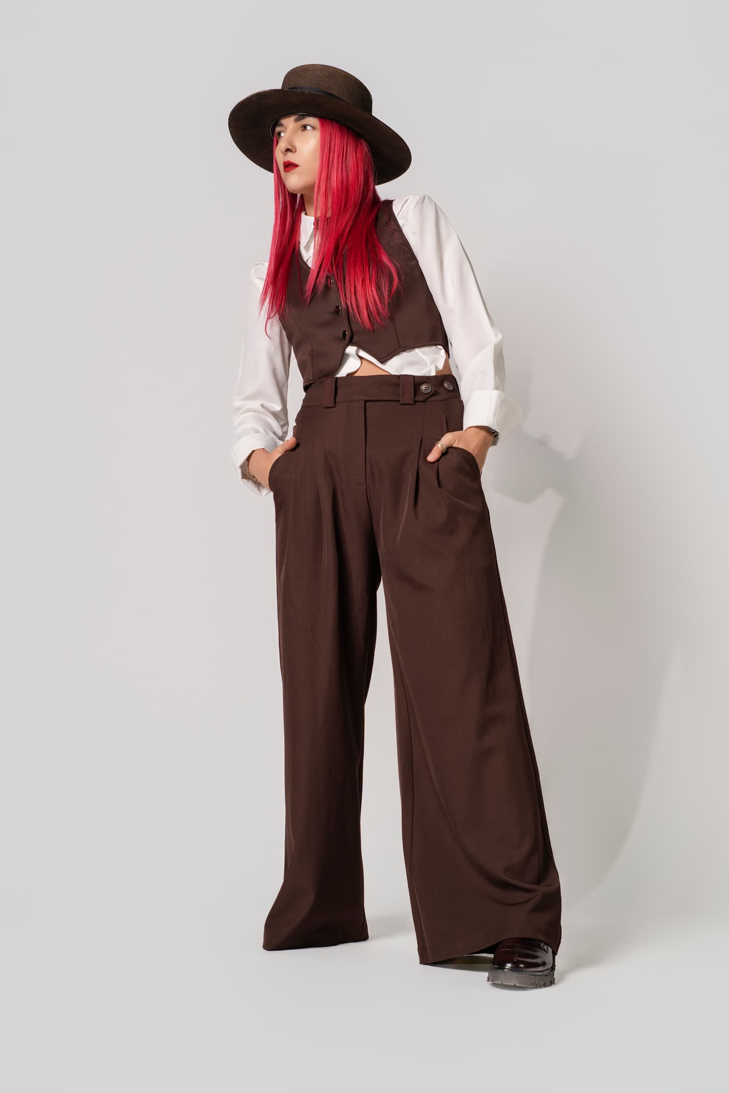 Modella con capelli rossi, cappello marrone, gilet e pantaloni larghi su sfondo grigio neutro.