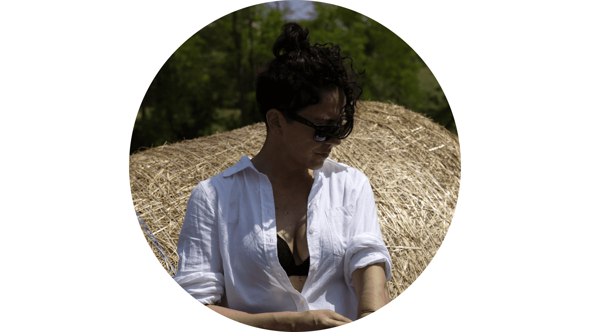 Donna con occhiali da sole e camicia bianca davanti a una balla di fieno.