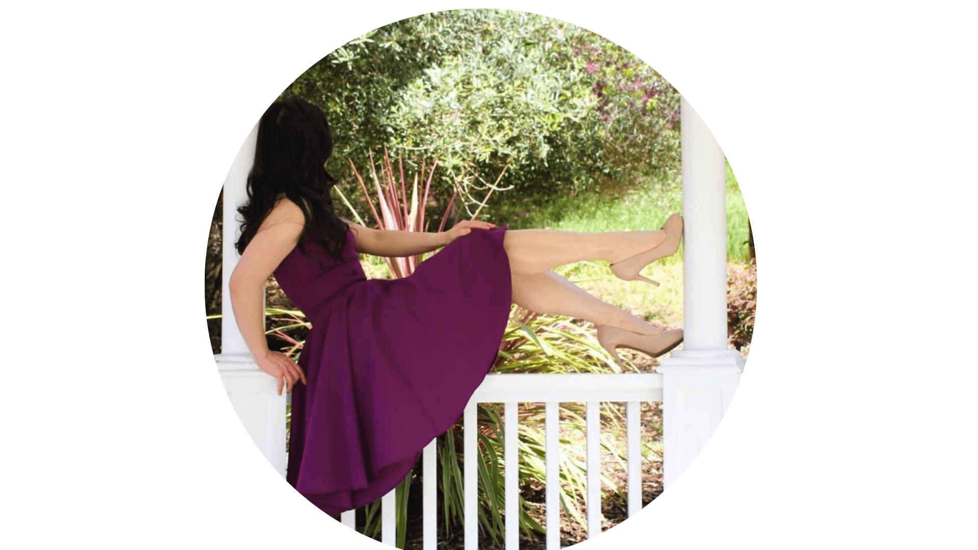 Donna in abito viola seduta su ringhiera bianca con gambe alzate e tacchi beige.