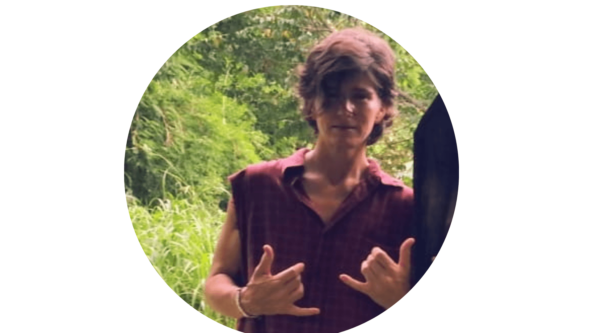 Persona con camicia a quadri bordeaux senza maniche fa il gesto shaka nella natura.