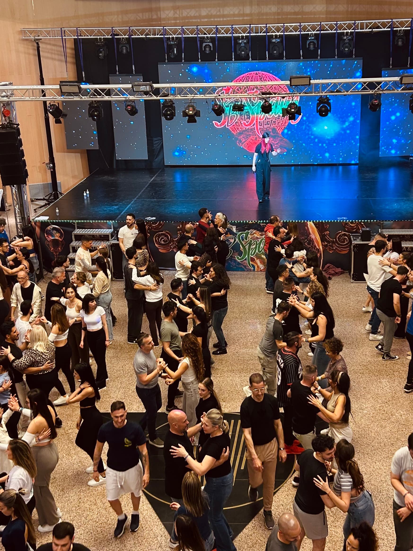 Numerose coppie ballano in una sala affollata durante un evento di danza con un palco.