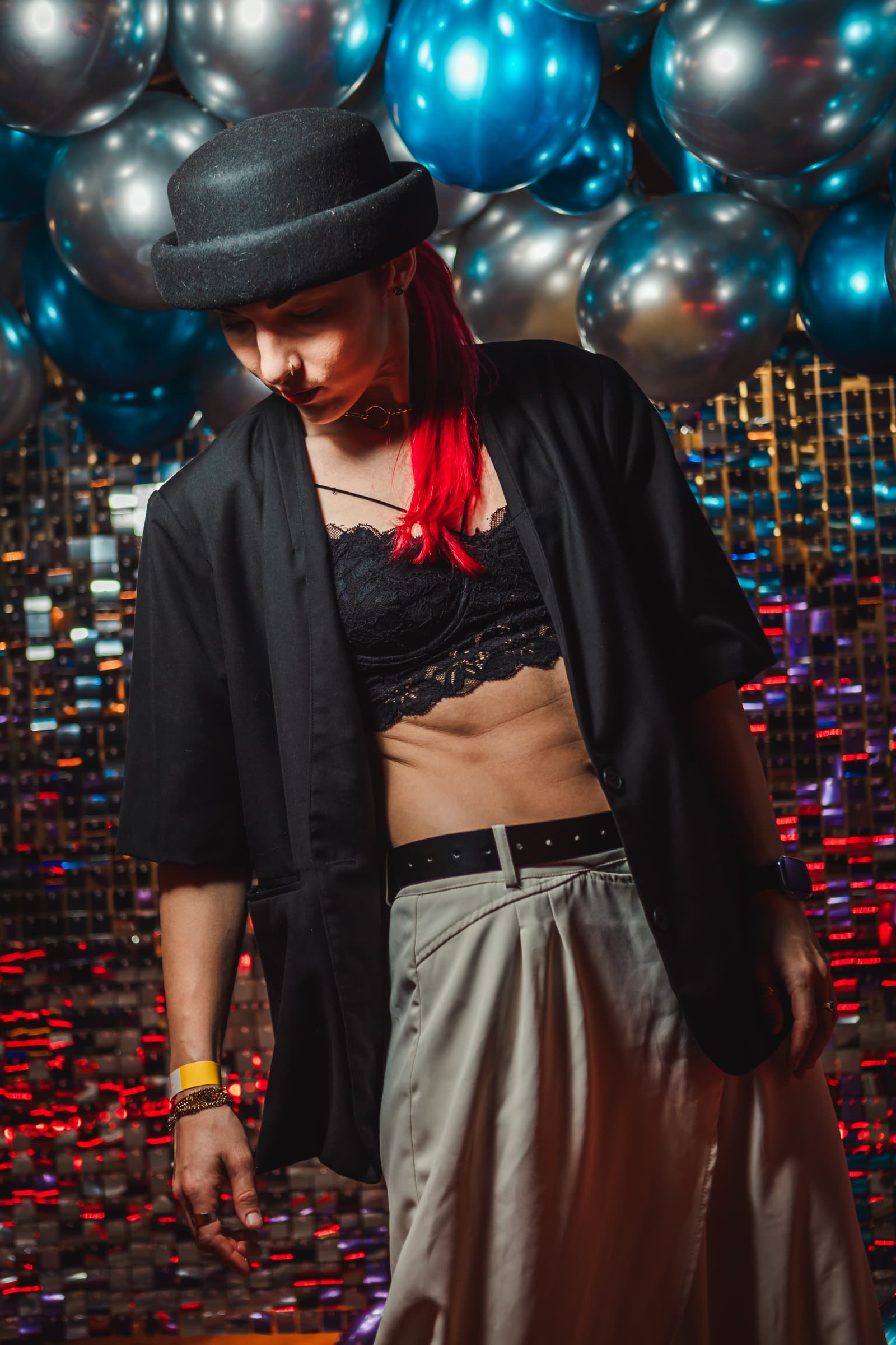 Persona con capelli rossi e bombetta nera davanti a palloncini blu e sfondo luccicante.