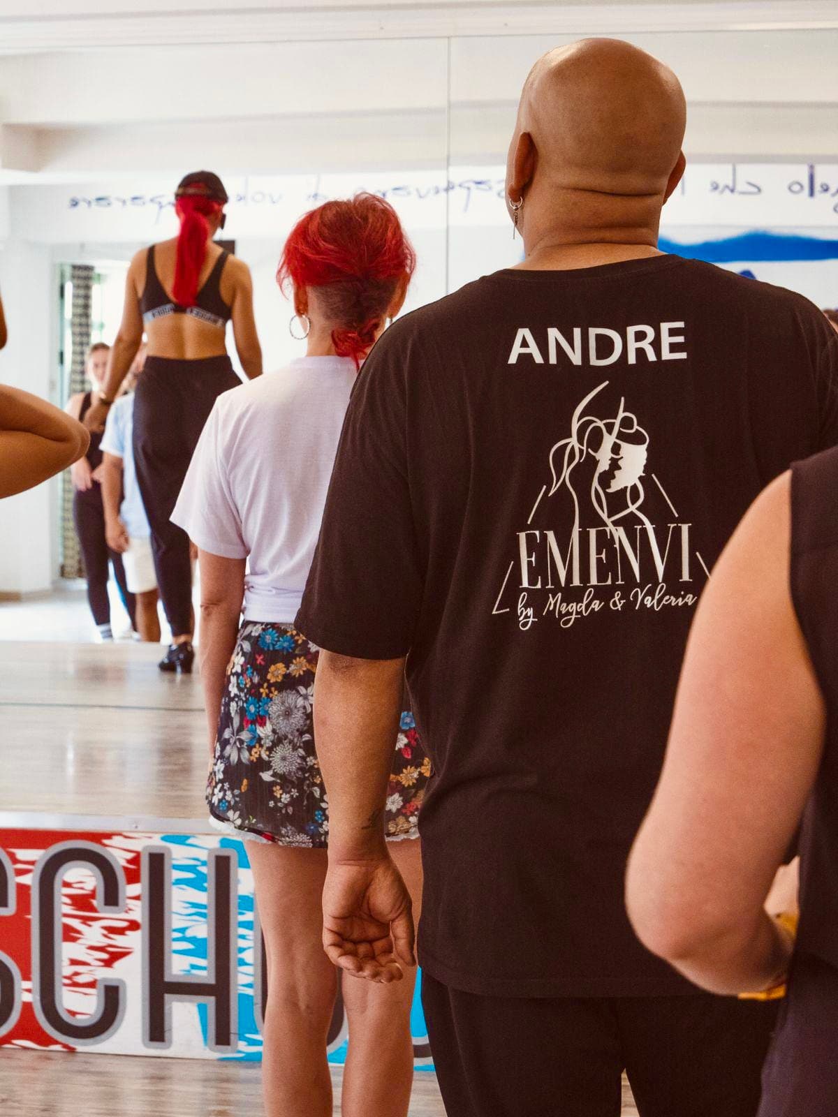 Ballerini in fila in una sala prove, uomo calvo in primo piano di spalle.